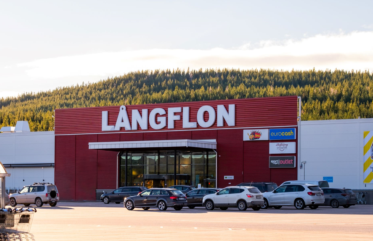 Långflon shoppingcenters fasad och parkering en solig dag. I bakgrunden står den vackra granskogen på ett fjäll. 